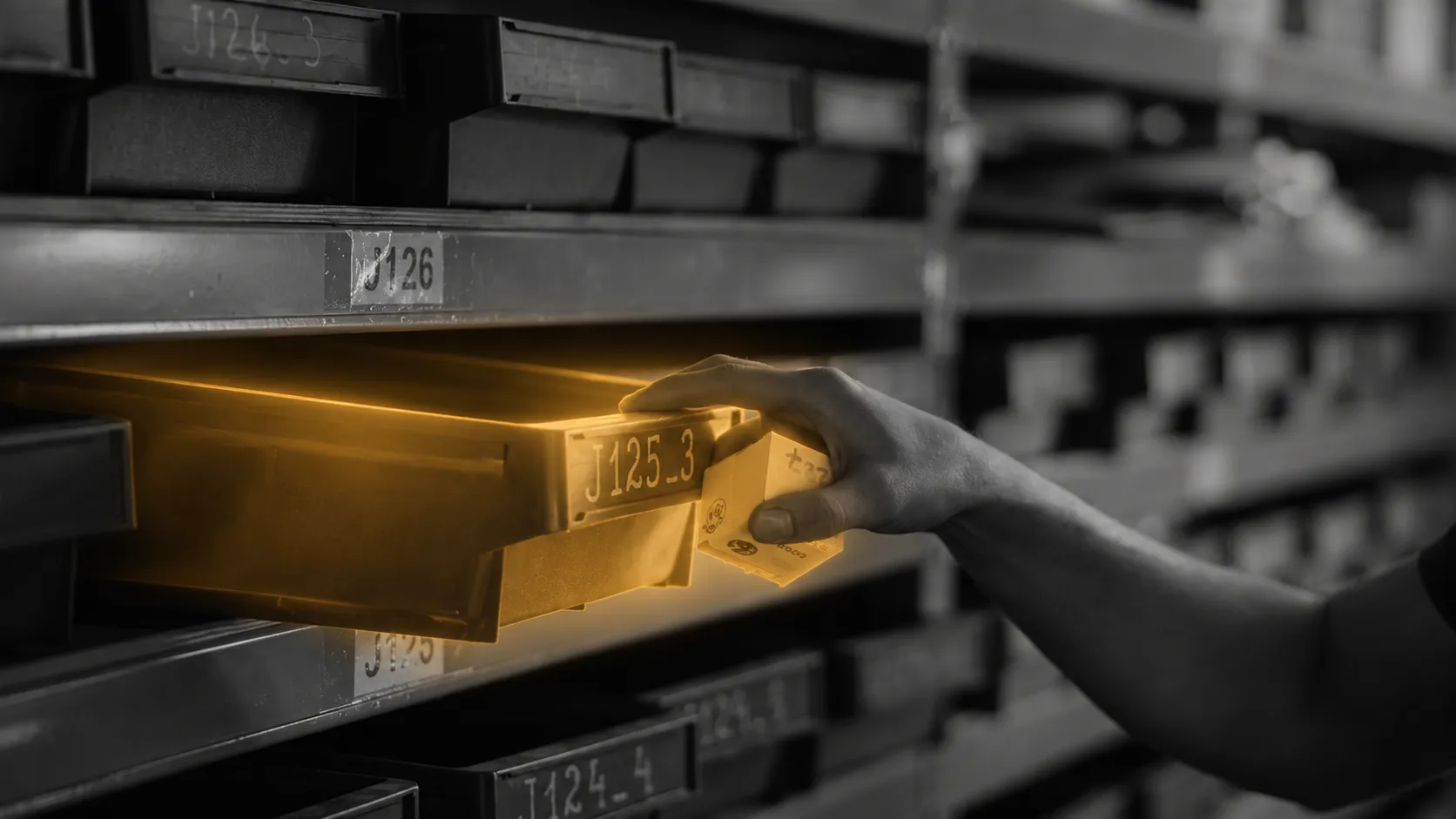 A hand pulls an amber-illuminated parts bin from industrial shelving.